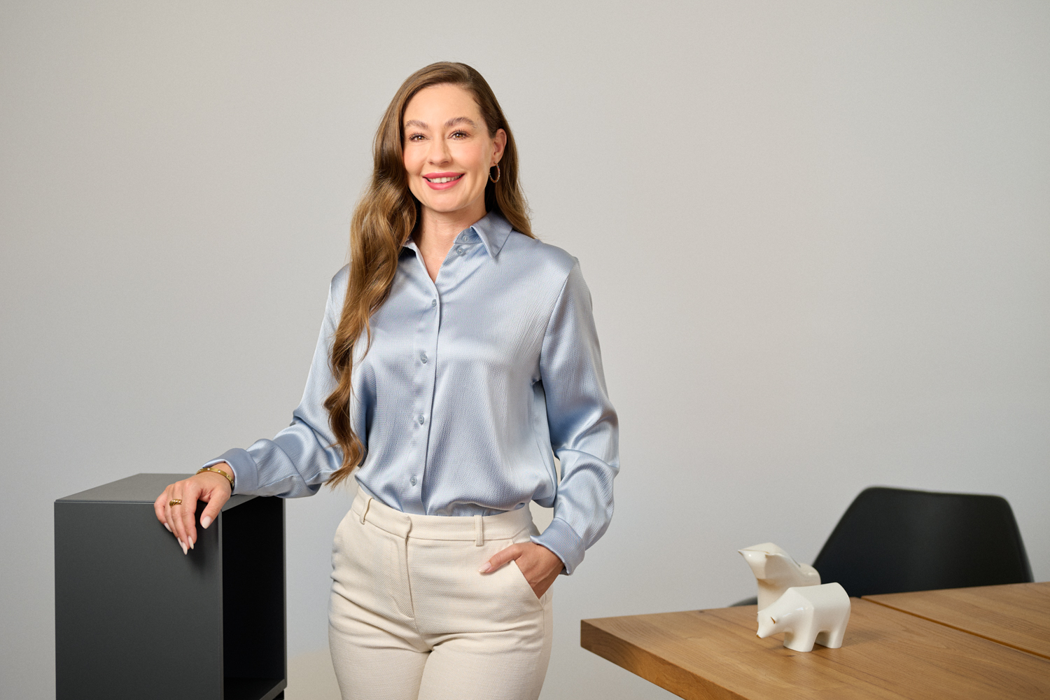 Portrait von Sarah Diersch: eine junge Frau mit langen, blonden Haaren, gekleidet mit einer hellblauer Bluse und einer weißen Hose, lehnt mit einer Hand an einem dunklen Möbelstück in einem modernen Büro. Auf dem Tisch im Hintergrund stehen weiße Stier- und Bärfiguren.