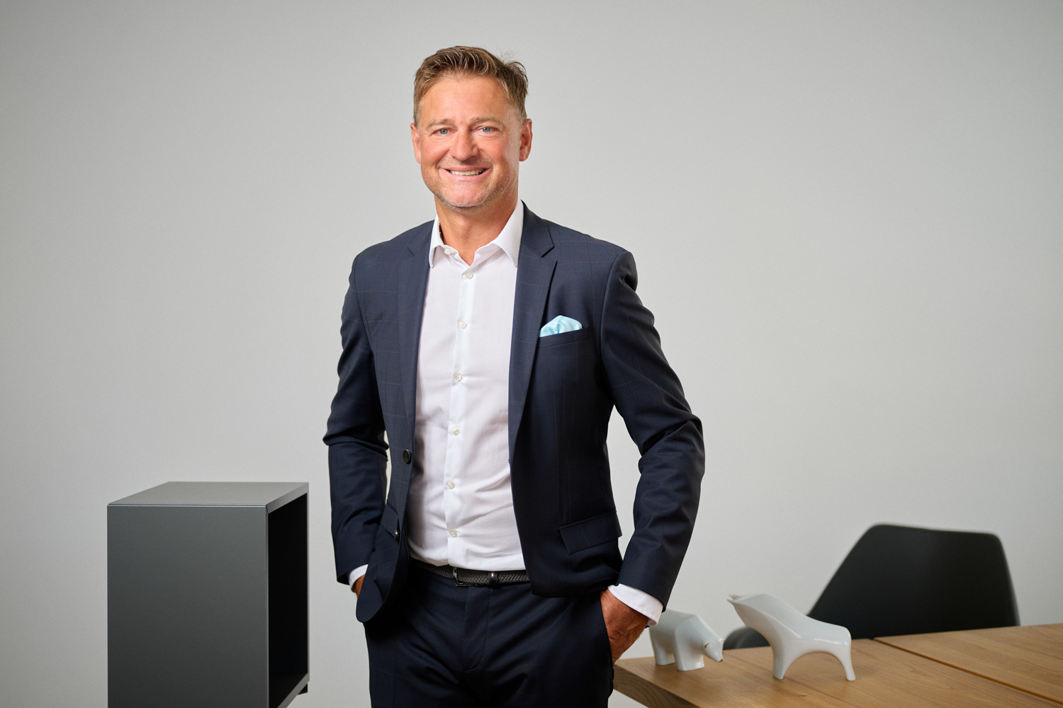 Portrait von Thomas Görnandt: ein Mann mittleren Alters mit kurzem, braunen Haar, dunklem Anzug und weißem Hemd lächelt in die Kamera. Er steht in einem modernen Büro, auf dem Tisch im Hintergrund stehen weiße Stier- und Bärfiguren.